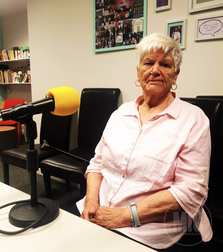 a photo of a woman sitting in a chair and looking at the camera. the woman is white, with white hair. on the table in front of her is a microphone with a yellow foam cover. The microphone is pointing towards her.