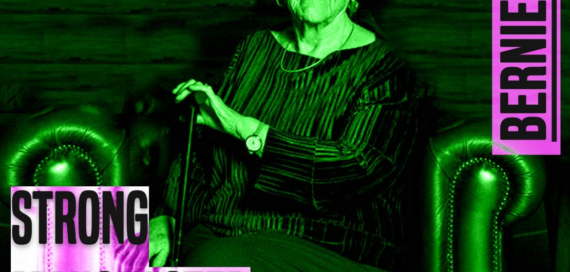 A photo of women sat in a leather Chesterfield style chair. The image is from her knees up. She is holding a walking stick. She is white. she is looking directly at the camera. She is wearing dark clothes. She has short white hair. The image has a green hue to it. to the left of her knees are the words 'strong manchester women' in grey lettering and a thick font in capital letters. In the same style font, next to her right ear are the words 'Bernie Wood'.