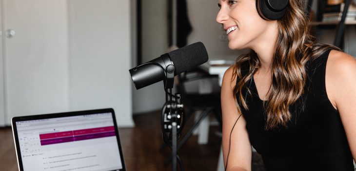 Image of a woman with headphones on and black vest top and microphone with a laptop in the background