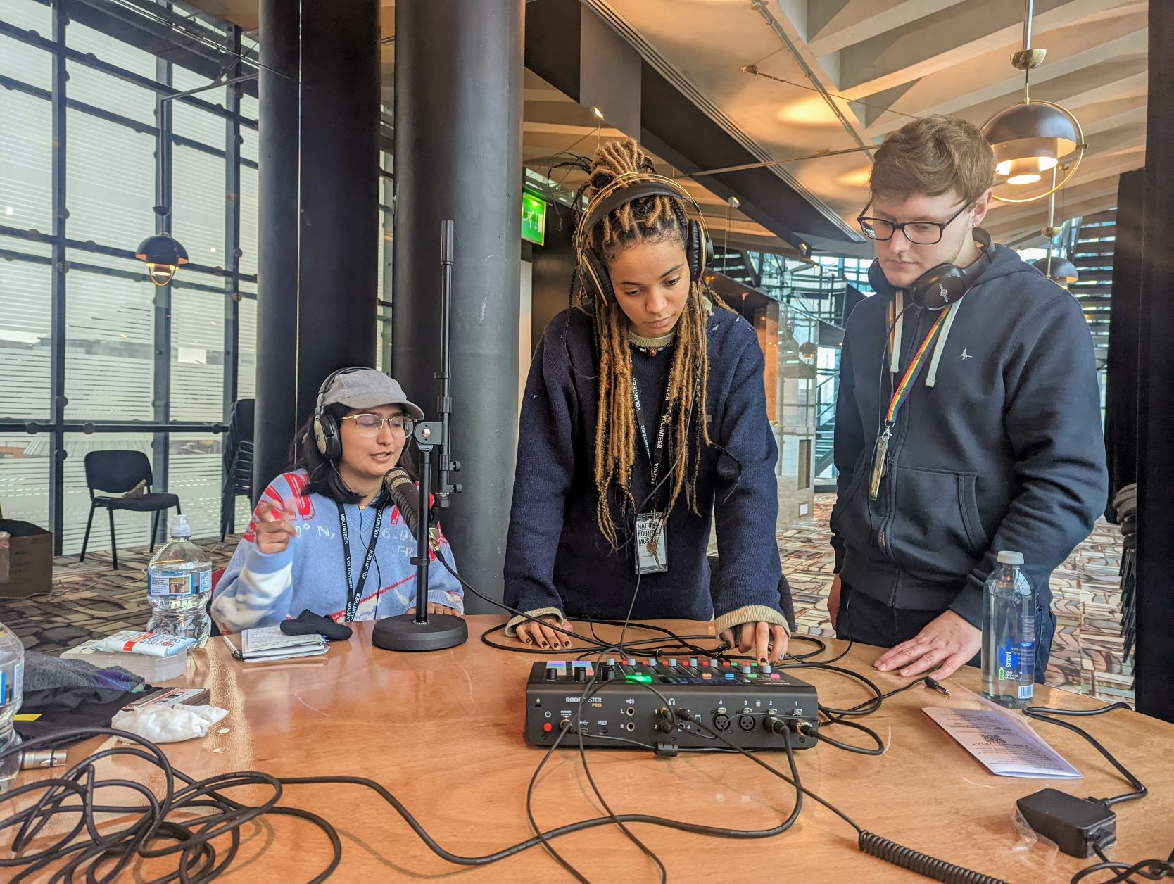 3 people around a table. 1 girl seated with headphones on. A male and young girl stand looking over a sound desk. The record the national football museum podcast