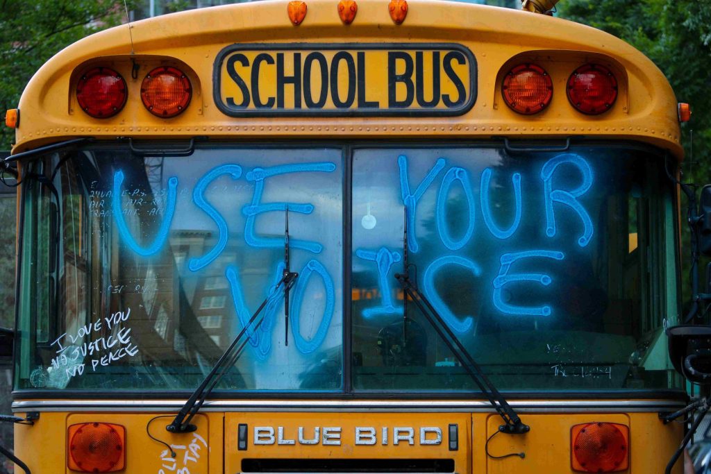 a yellow school bus with 'use your voice' spraypainted blue across the windscreen