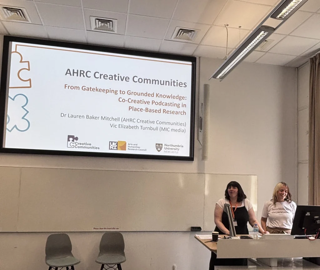 two women present in fron of a laurge screen that says AHRC Creative Communities on it