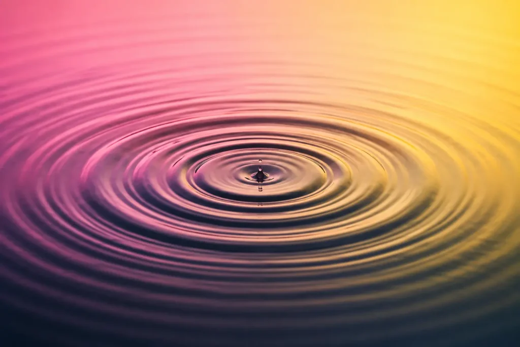 Concentric ripples spreading across calm water with soft pink and yellow reflections.